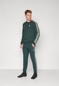Dunkelgrüner Trainingsanzug mit einem Sweatshirt mit Rundhalsausschnitt, weißen Streifen an den Ärmeln und passenden Hosen, beide mit goldenen Logo-Details. Schwarze Schuhe.