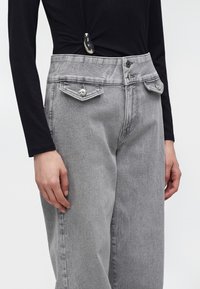 Personne portant un jean gris taille haute avec poches à rabat devant et fermeture double bouton, associé à un haut noir à manches longues.
