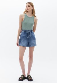 Grön linne topp kombinerad med ljusblå denimshorts med dragsko i midjan, och svarta sandaler med två spännen. En enkel vit bakgrund.