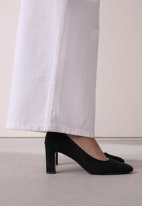 Black suede high heels with a pointed toe design and a chunky heel, paired with white wide-leg trousers. Smooth texture, sleek silhouette.