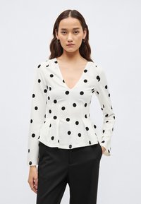 Hvit genser med lange ermer og V-hals, med svarte polkadotter og peplumkant. Materialet virker glatt med en svak glans.