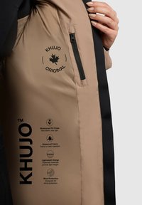 Beige pufferjack met zwarte accenten, voorzien van een ritszak, waterdichte stof en bedrukte verzorgingsinstructies voor warmte en bescherming.