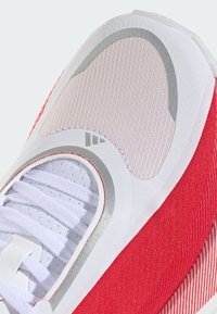 Gros plan sur une chaussure de sport blanche et rouge avec des lacets blancs, un tissu en mesh et des détails argentés réfléchissants autour de la zone des orteils et des lacets.