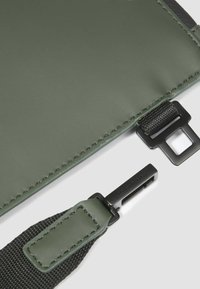 Olijfgroene clutch met een gladde textuur, voorzien van een zwarte riem, kunststof hardware en gestikte randen voor extra detail.