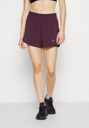 Woman wearing maroon Adidas athletic shorts, black sports bra, and black running shoes, standing against a plain light background.