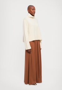 Dorosła osoba w kremowym swetrze z golfem, szerokich brązowych spodniach plisowanych i butach w panterkę, stojąca na tle jednolitego białego tła.