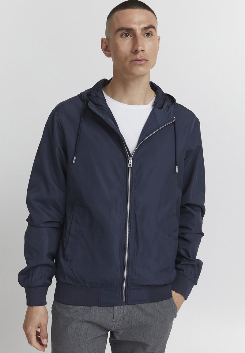 Marineblauwe lichtgewicht jack met een capuchon, ritssluiting aan de voorkant, elastische manchetten en tailleband. Glad textuur met zijzakken en koorddetails.