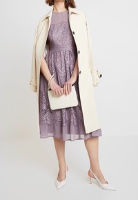 Ljuslila spetsklänning med figurnära överdel och utsvängd kjol, i kombination med en beige kappa och vit handväska. Modellen bär vita spetsiga klackar.