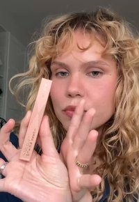 Personne aux cheveux bouclés tenant une boîte rectangulaire rose de gel à sourcils étiquetée "CURL'DS ARROW" près de son visage à l'intérieur.