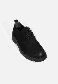 Chaussure à lacets en suède noir avec un bout rond, dotée d'une semelle en caoutchouc épaisse et texturée, et d'un design minimaliste.