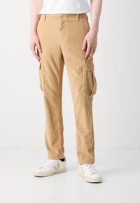 Beige corduroy cargo-housut, joissa on sivutaskut, yhdistetty valkoisiin lenkkareihin ja valkoiseen paitaan, seisoo valkoista taustaa vasten.