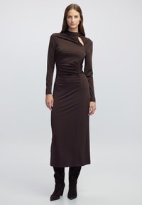 Braunes langärmeliges Kleid mit hohem Kragen, das seitliche Raffung und einen Schlüsselloch-Ausschnitt aufweist. Der Stoff wirkt glatt und körperbetont.