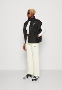 Černá prošívaná vesta s vysokým límcem, logo Nike na levé části hrudi, spárována s bílým dlouhým rukávem a krémovými širokými kalhotami, černé tenisky.