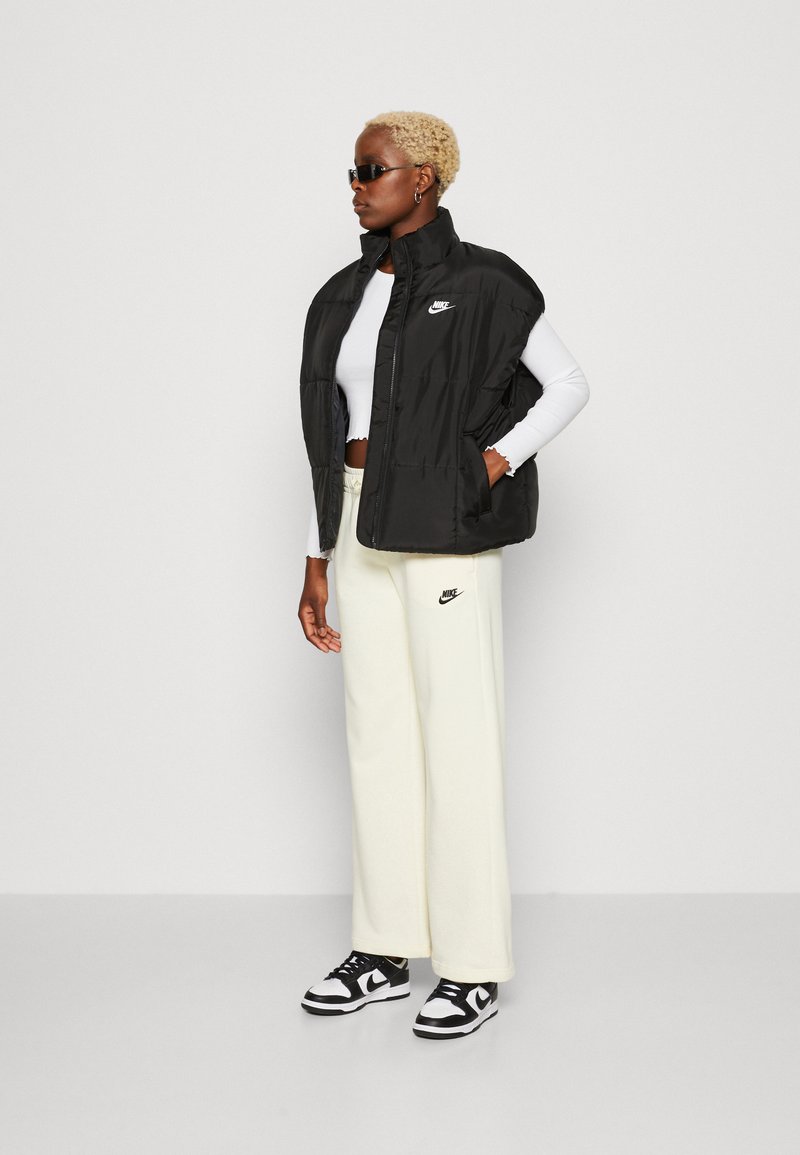 Black padded vest with high collar, Nike logo on left chest, paired with a white long-sleeve top and cream wide-leg pants, black sneakers.