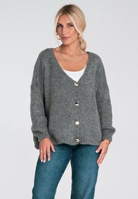 Szary kardigan dzianinowy z głębokim dekoltem w kształcie V, oversize'owymi rękawami, pięcioma dużymi guzikami i ściągaczem na dole, noszony na białej bluzce i niebieskich jeansach.