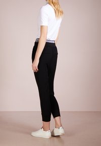 Kobieta w czarnych dresowych spodniach z logo w talii, białej koszulce z krótkim rękawem i białych trampkach z metalicznymi srebrnymi akcentami na pięcie, stojąca bokiem.