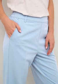 Donna che indossa pantaloni azzurri chiari e una maglietta bianca, con la mano sinistra in tasca e la mano destra appoggiata sulla coscia, mostrando unghie curate.