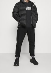 Man som bär svart puffjacka, svart grafiktröja, svarta uppvikta byxor, vita strumpor och svarta sneakers, står med händerna i fickorna.