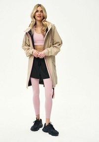 Kobieta stojąca w beżowej, długiej kurtce z kapturem, nałożonej na różowy sportowy biustonosz, czarne szorty, różowe legginsy i czarne trampki na białym tle.