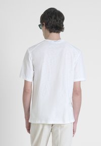 Maglietta bianca a maniche corte con colletto rotondo, texture liscia e una leggera lucentezza, indossata con pantaloni chiari e larghi.