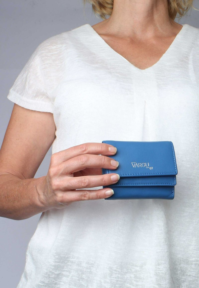 Blue leather wallet with two compartments, featuring stitching details and a metallic logo. Held by a hand, against a white top.