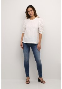 Top bianco texturizzato con maniche a palloncino e scollatura rotonda, abbinato a jeans blu aderenti e sandali neri con cinturini su uno sfondo neutro.