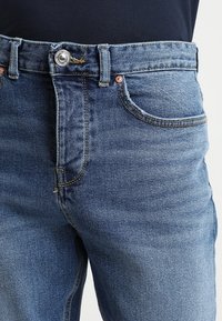 Vaqueros de mezclilla en azul medio, de talle alto con un diseño clásico de cinco bolsillos, con un botón metálico y detalles de costura sutiles.