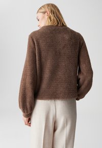Brauner flauschiger Pullover mit kurzem Schnitt und Ballonärmeln, getragen über hellen Hosen, der eine weiche Textur zeigt.