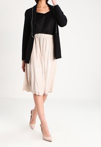 Nainen, joka on pukeutunut mustaan bleiseriin, mustaan toppiin, beigevalkoiseen, värikäs midi-hameeseen ja beigeihin korkokenkiin, seisoo jalat ristissä.