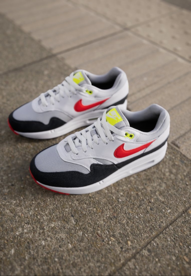 Une paire de baskets Nike Air Max blanches, noires et grises avec des logos swoosh rouges et des accents jaunes néon, posées sur un pavé texturé.