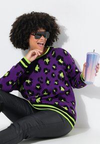 Maglione a maglia viola con stampa leopardata, accenti verde lime e strisce nere. Indossato con pantaloni in pelle nera, mentre tiene in mano una tazza olografica testurizzata.