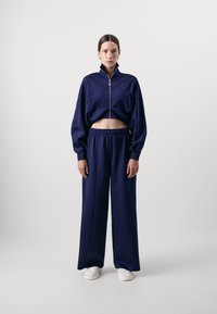 Giacca corta zip-up blu navy con dettagli di cuciture verticali e pantaloni a gamba larga coordinati. Tessuto morbido con vita elasticizzata, abbinata a sneakers bianche.