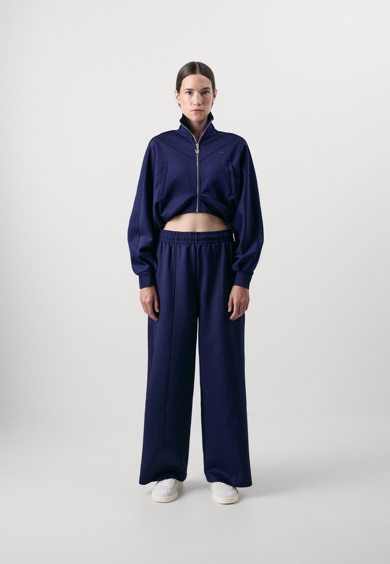 Giacca corta zip-up blu navy con dettagli di cuciture verticali e pantaloni a gamba larga coordinati. Tessuto morbido con vita elasticizzata, abbinata a sneakers bianche.