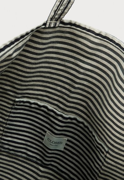 Εσωτερικό τσάντας tote από υφασμάτινο ριγέ, με εναλλασσόμενες λεπτές μαύρες και κρεμ ρίγες, και μια ετικέτα με την ένδειξη «CLOSED» ραμμένη στο πλάι.