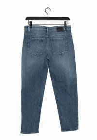 Blauwe denim jeans hangen aan een zwarte hanger; ze hebben een rechte pijp, vijf zakken en een leren patch op de tailleband.