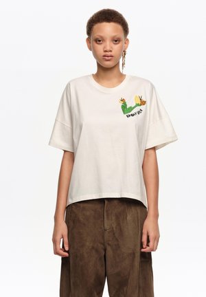 Persona joven con camiseta blanca con bordado de caracol verde y amarillo y pantalones holgados marrones, de pie contra un fondo blanco.