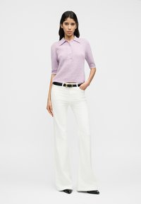 Γυναίκα στέκεται με το χέρι στην τσέπη φορώντας ανοιχτό μωβ πλεκτό πόλο, λευκά φαρδιά παντελόνια, μαύρη ζώνη και μαύρα μυτερά παπούτσια πάνω σε λευκό φόντο.