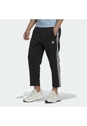 Mężczyzna ubrany w czarne spodnie dresowe Adidas z białymi paskami i białe trampki, stojący z jedną ręką w kieszeni na jednolitym tle.