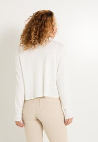 Haut blanc court à manches longues avec une texture douce. Présente un décolleté large et des manches légèrement évasées, associé à un jean beige taille haute.