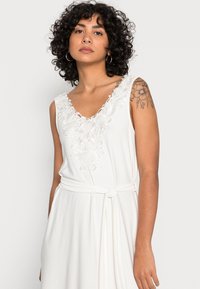 Vestido branco sem mangas com bordados florais ao redor da gola, com uma cintura amarrada e uma textura suave.