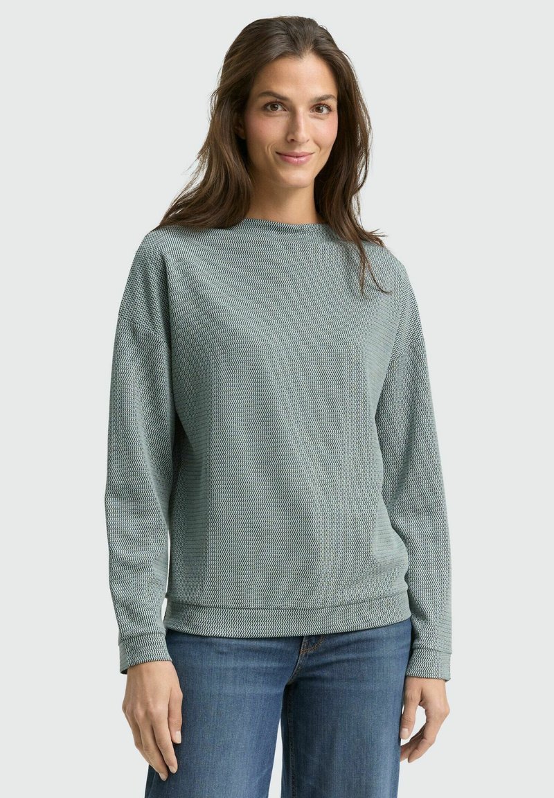 Light green patterned sweatshirt with long sleeves, round neckline, and ribbed cuffs; worn with blue jeans. Soft texture and relaxed fit.
