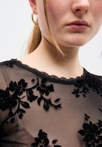 Μαύρο διάφανο τοπ με κεντήματα από βελούδο με λουλουδένιο σχέδιο. Διαθέτει ντεκολτέ με δαντελωτή λεπτομέρεια και κοντά μανίκια. Λεπτή υφή, εφαρμοστή γραμμή.