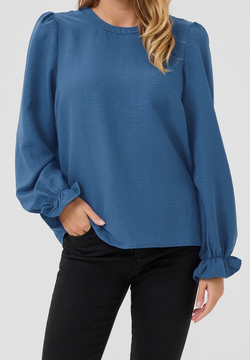 Femme portant un chemisier bleu à manches longues avec poignets froncés et un pantalon noir, une main dans la poche et les cheveux lâchés sur l'épaule.