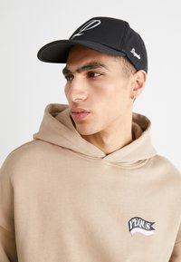 Black cap with embroidered logo, mesh sides; beige hoodie with graphic patch. Close-up of a person facing right, neutral background.