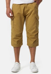 Shorts cargo jusqu'aux genoux en coton khaki avec poches latérales et coutures ton sur ton. Portés avec des chaussures de sport blanches.