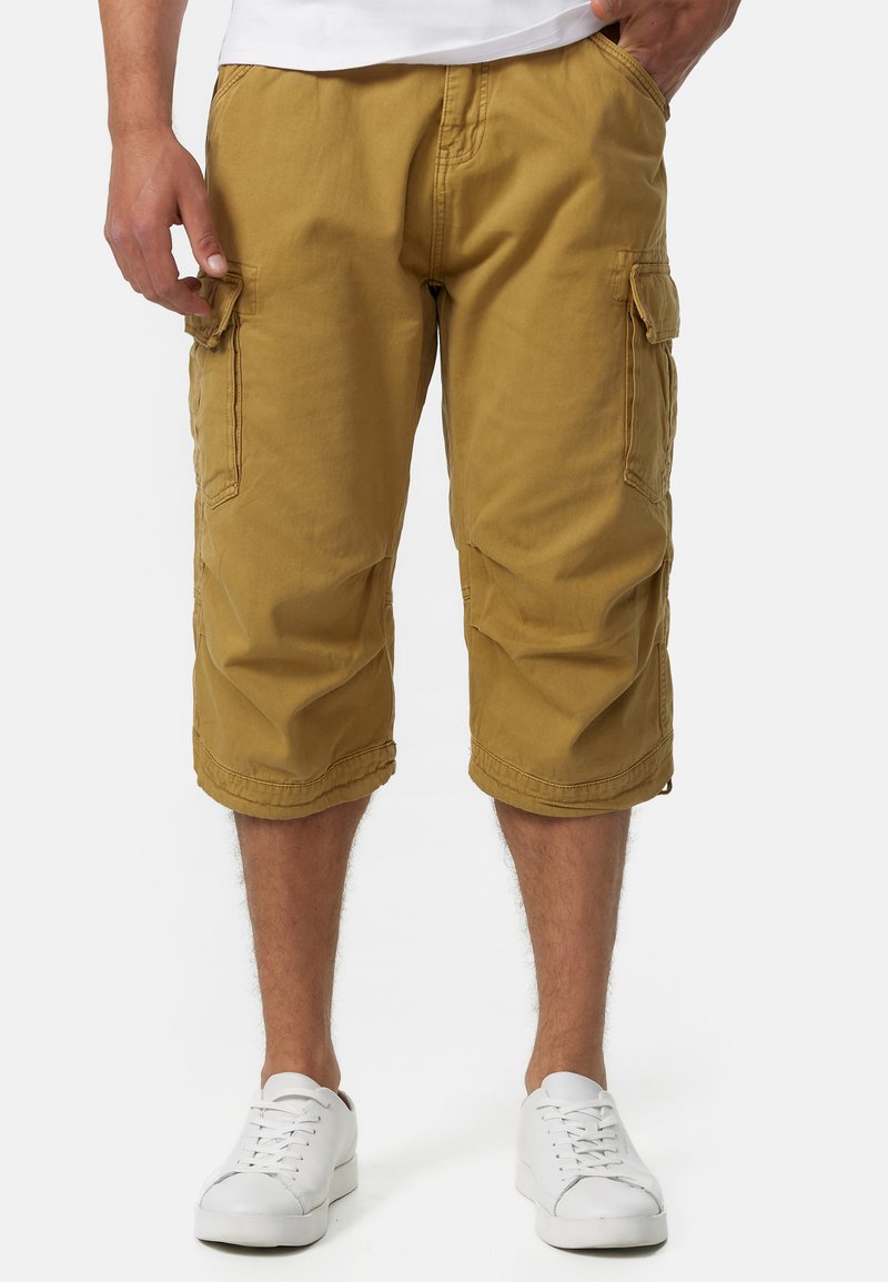 Shorts cargo jusqu'aux genoux en coton khaki avec poches latérales et coutures ton sur ton. Portés avec des chaussures de sport blanches.