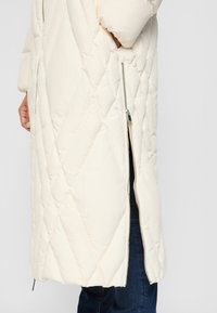 Long, cream puffer jacket featuring diamond quilting, a zipper on the side, and a relaxed fit. Worn over dark jeans.