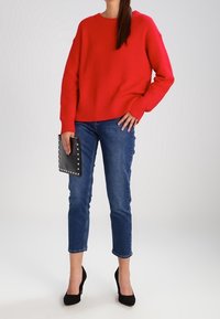 Red ribbed sweater with long sleeves, paired with blue denim jeans and black high heels. Holding a black studded clutch.