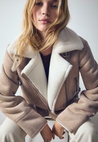 Mladá žena s blond vlasmi, oblečená v béžovej kožúškovej bunde s bielym kožúškom, sedí so založenými rukami.