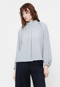 Hellgraue Bluse mit hohem Kragen, Falten-Details und langen Ballonärmeln, die an den Bündchen gerafft sind. Glatter, fließender Stoff.